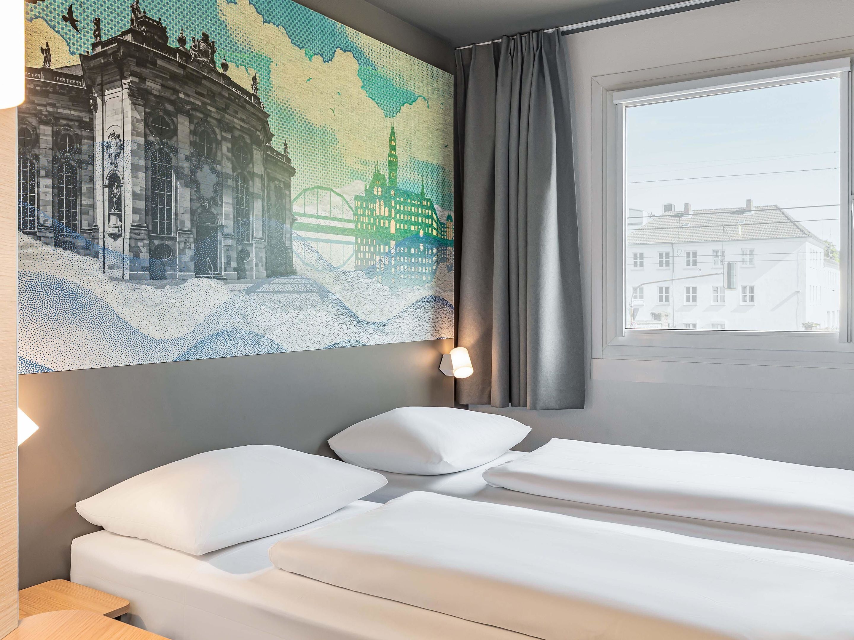 A hotel room with a bed, a wall-mounted artwork of a cityscape, a window with curtains, and a view of buildings outside.