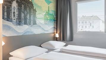 A hotel room with a bed, a wall-mounted artwork of a cityscape, a window with curtains, and a view of buildings outside.