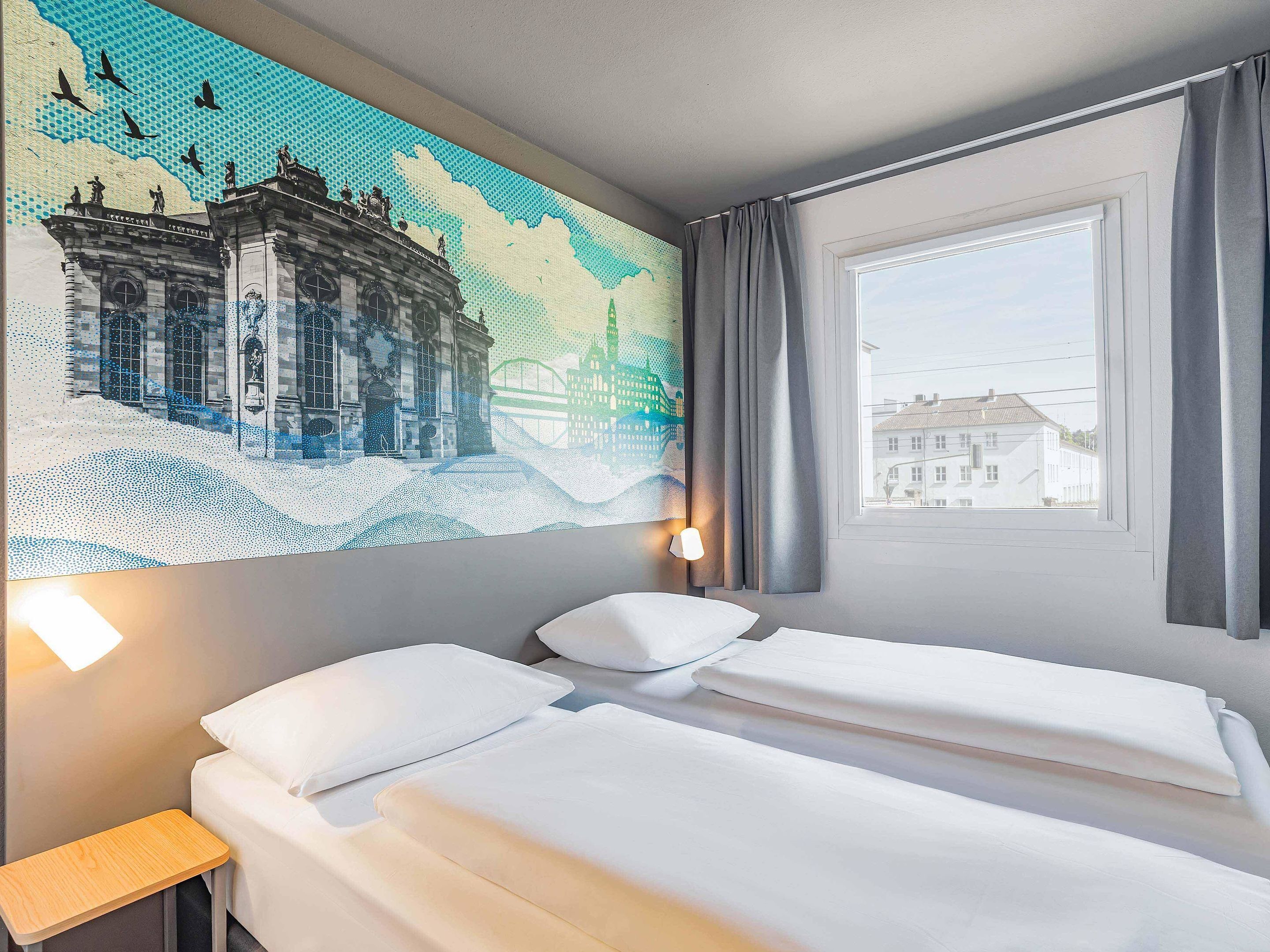 Ein Hotelzimmer mit zwei Betten, einem Nachttisch, einer Wandlampe und einem Fenster mit Blick auf Gebäude.