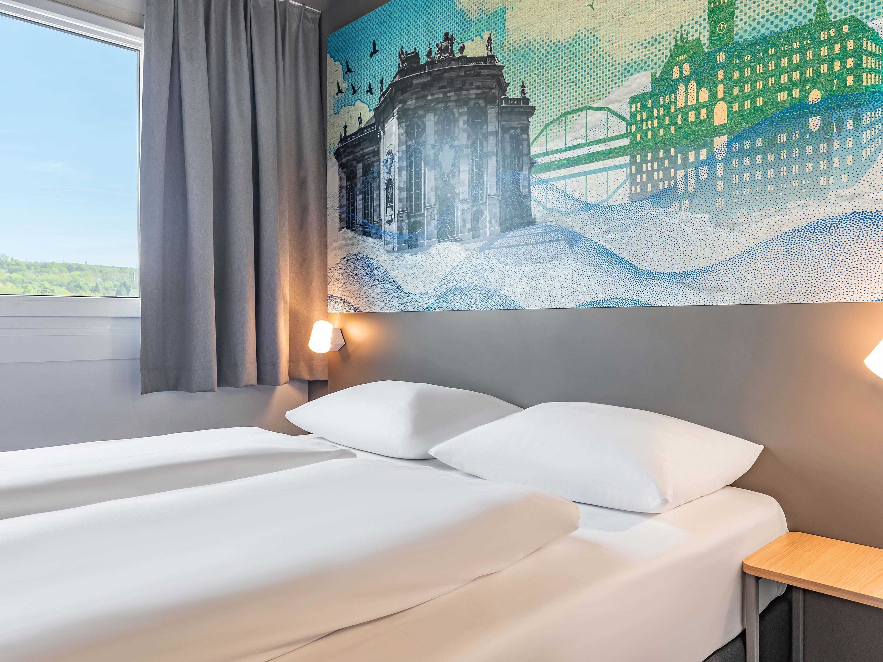 A hotel room with a bed, a nightstand, a wall-mounted lamp, and a large window with curtains.