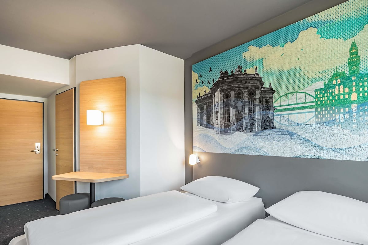 Une chambre d’hôtel avec deux lits, un bureau en bois, une lampe fixée au mur et un grand tableau représentant un paysage urbain.
