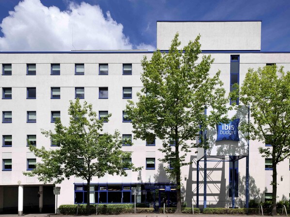 Een modern gebouw met veel ramen, een blauw bord met 'Ibis Budget' en een boom op de voorgrond.