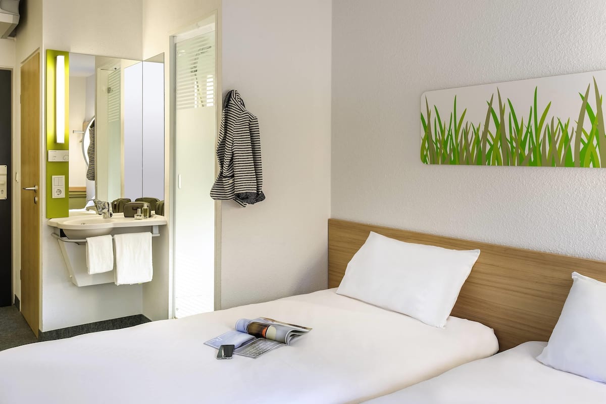 Habitación de hotel con cama, mesita de noche, espejo, baño con lavamanos y toallero, y una pared decorada con una planta verde.