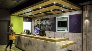 A reception desk with a counter, a woman in a blue uniform, and a man walking by. - Ibis Budget Nuernberg City MES (Nuremberg)