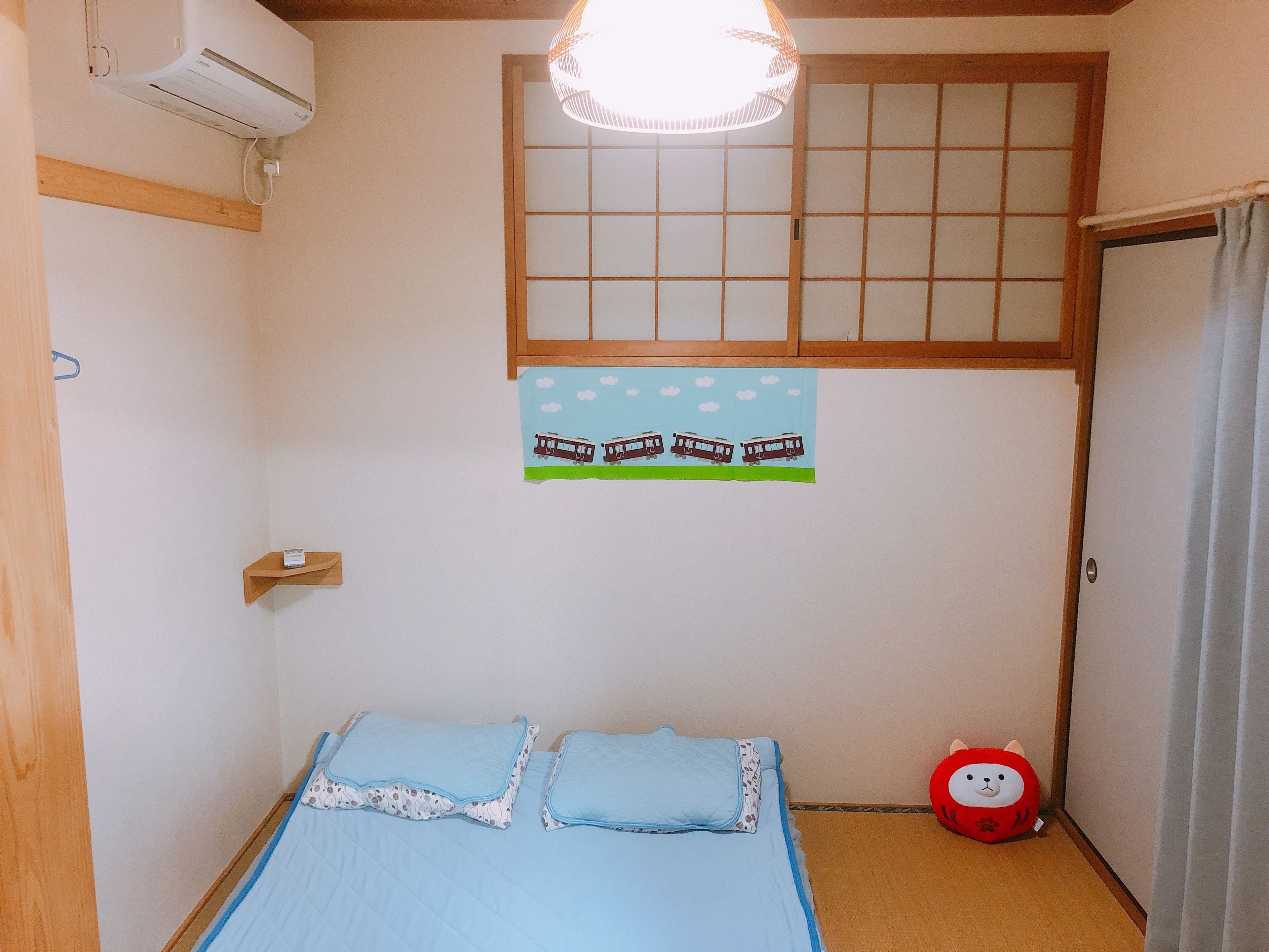 A bedroom with a bed, a window with a view of the sky, a wall-mounted air conditioner, and a small shelf.