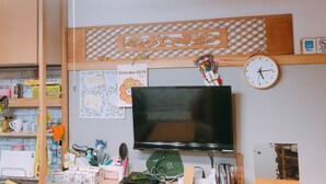 A room with a flat-screen TV, a wall clock, a calendar, and various items on shelves and a desk.