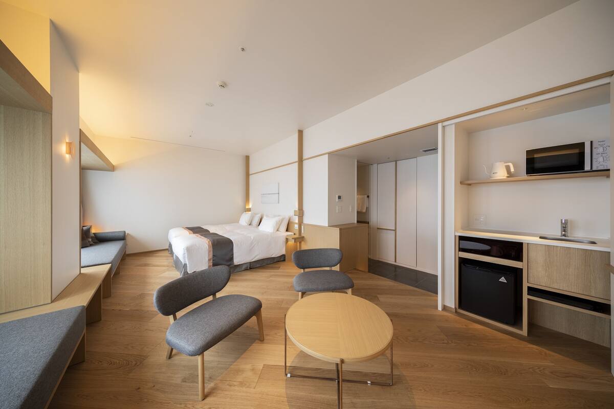A modern hotel room with a bed, seating area, and kitchenette.
