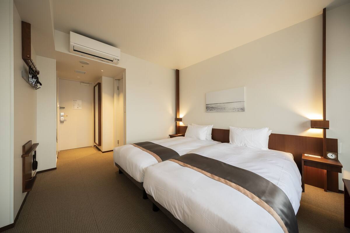 A hotel room with two beds, a wooden headboard, a nightstand with a clock, a wall-mounted air conditioner, and a framed picture on the wall.