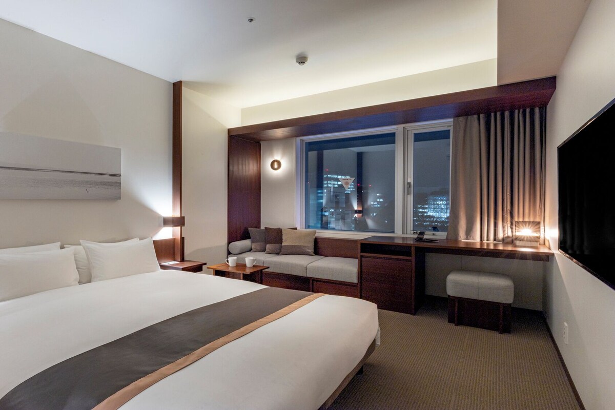 A hotel room with a large bed, a desk, a sofa, and a view of the city at night.