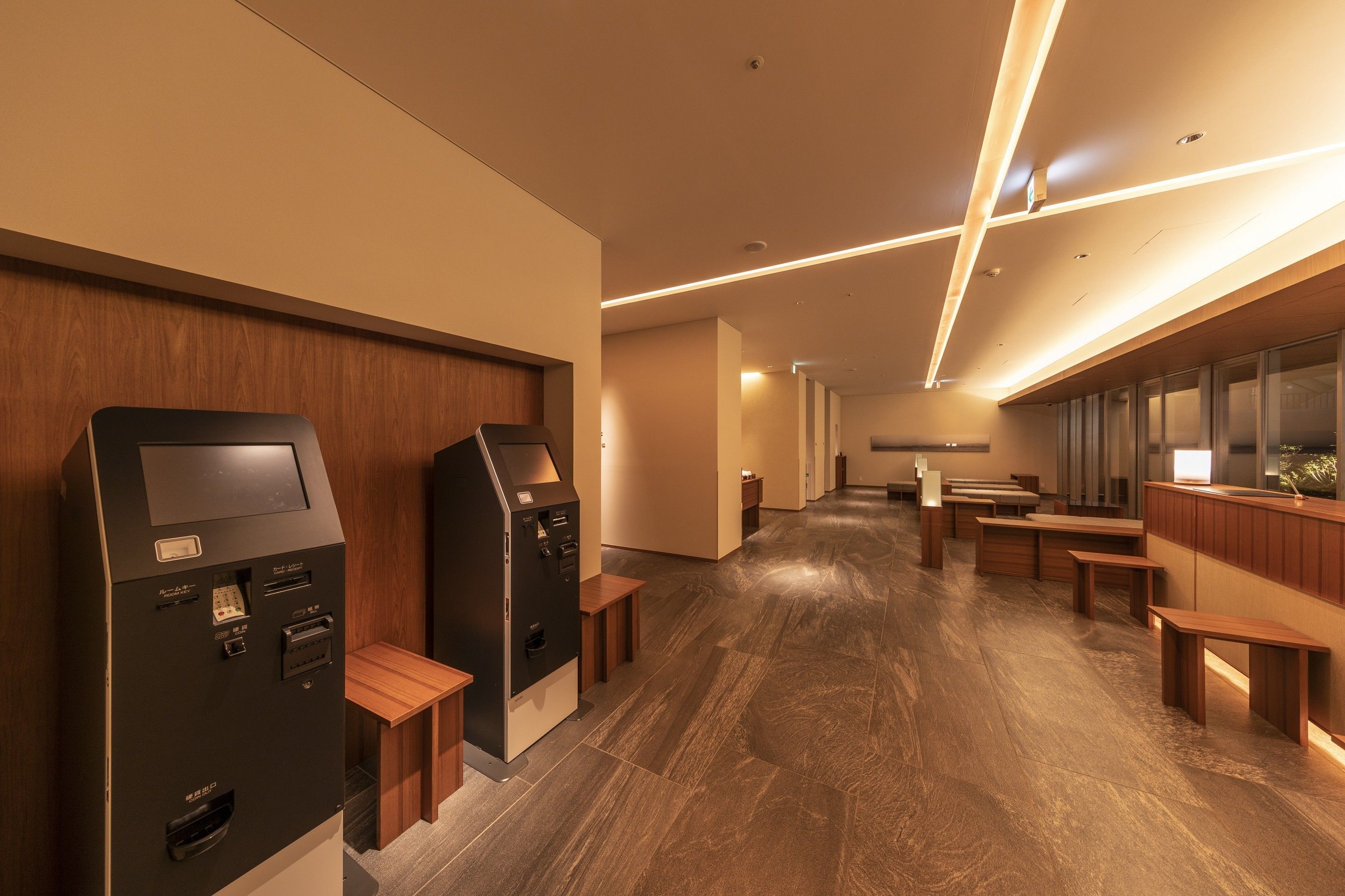 A modern hotel lobby with a wooden floor, two self-service kiosks, and a seating area with wooden benches.