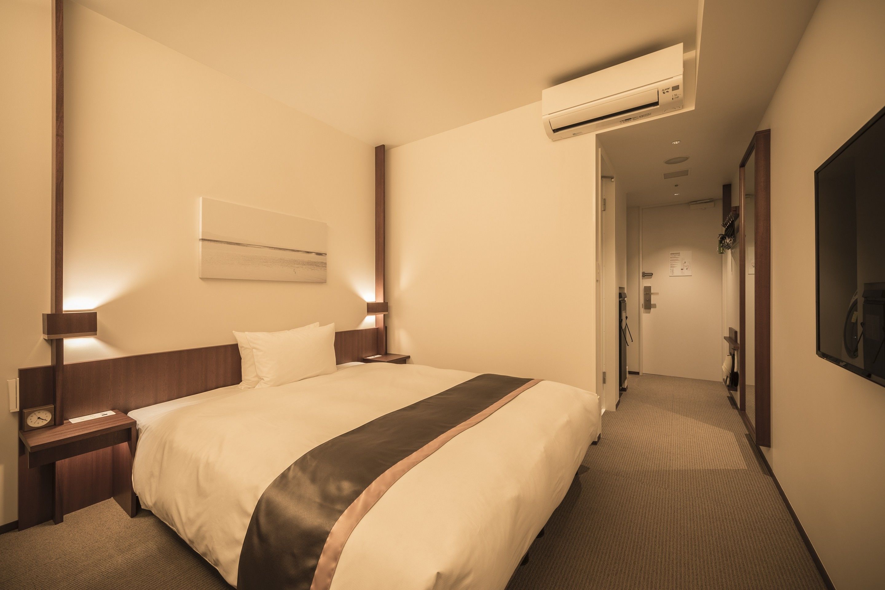 A hotel room with a large bed, bedside tables, an air conditioning unit, and a hallway leading to a door.