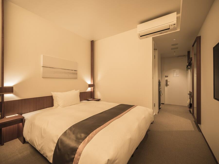 Habitación de hotel con una cama grande, mesitas de noche, un equipo de aire acondicionado y un pasillo que conduce a una puerta.