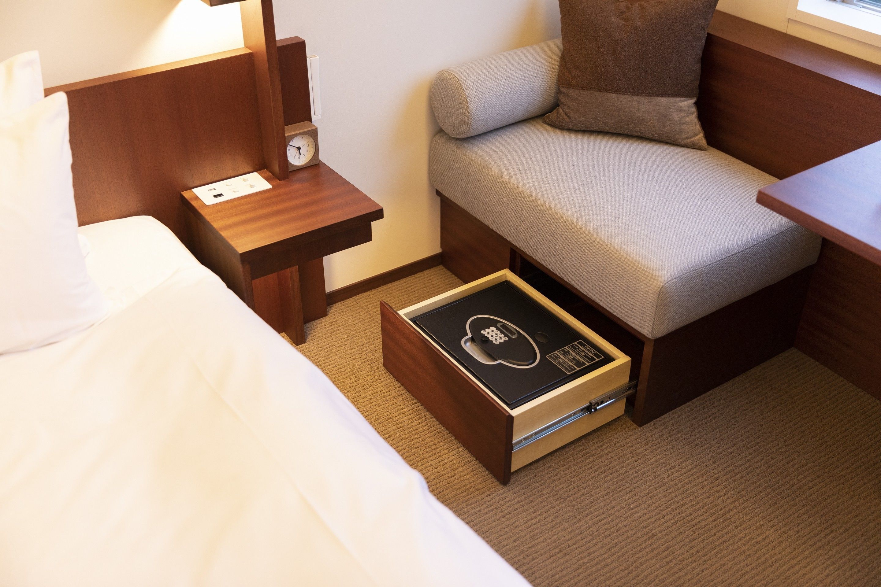 A compact hotel room with a bed, a small bedside table with a lamp, a sofa, and a drawer unit with a control panel.