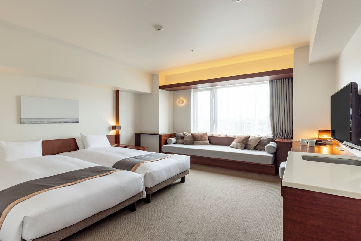 A hotel room with two beds, a desk, a TV, and a large window with curtains.