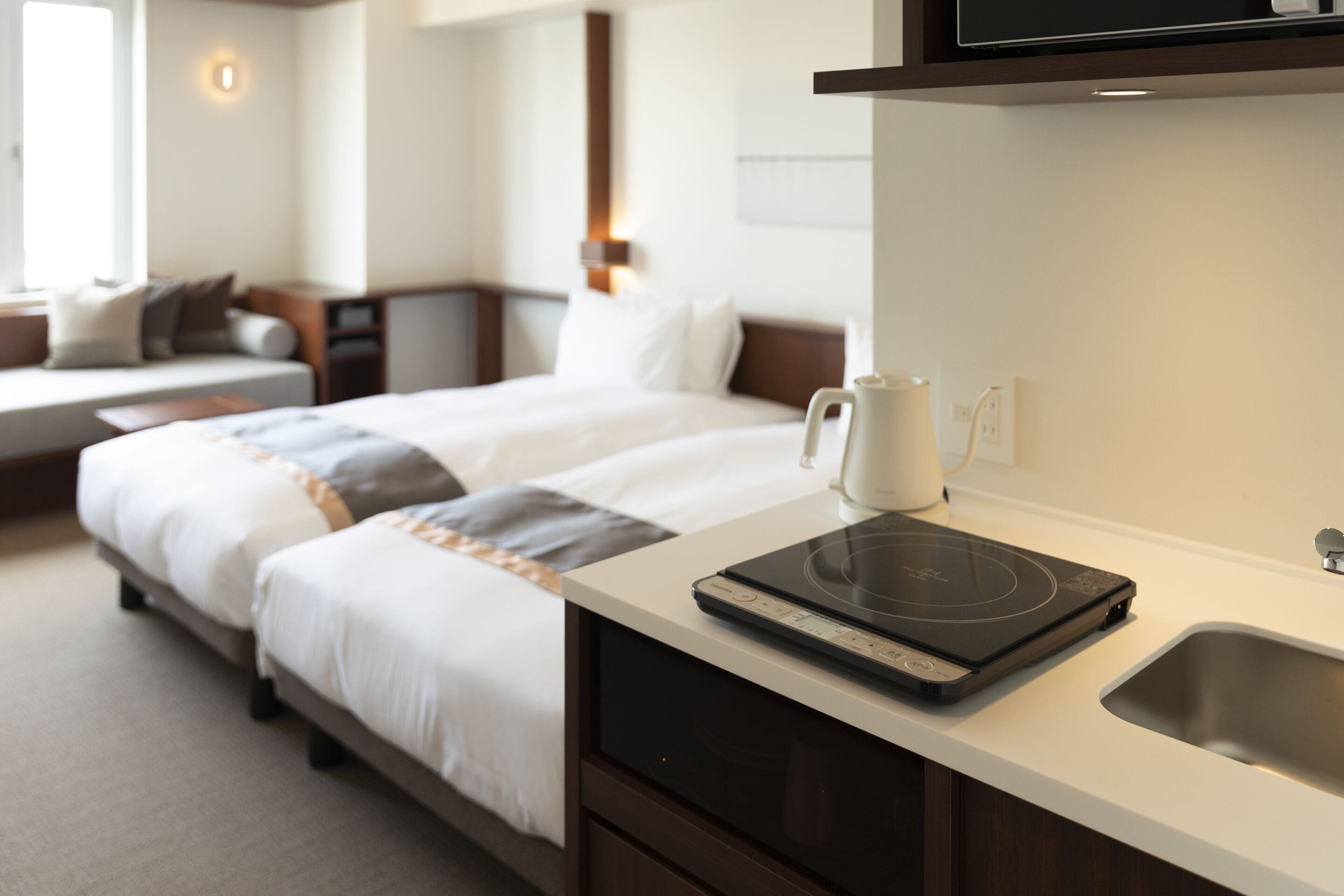 A hotel room with a bed, a coffee maker, a kettle, and a built-in induction cooktop on the counter.