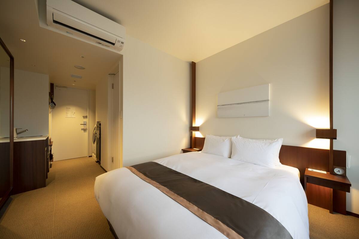 A hotel room with a large bed, bedside tables, a wall-mounted air conditioner, and a door leading to a bathroom.