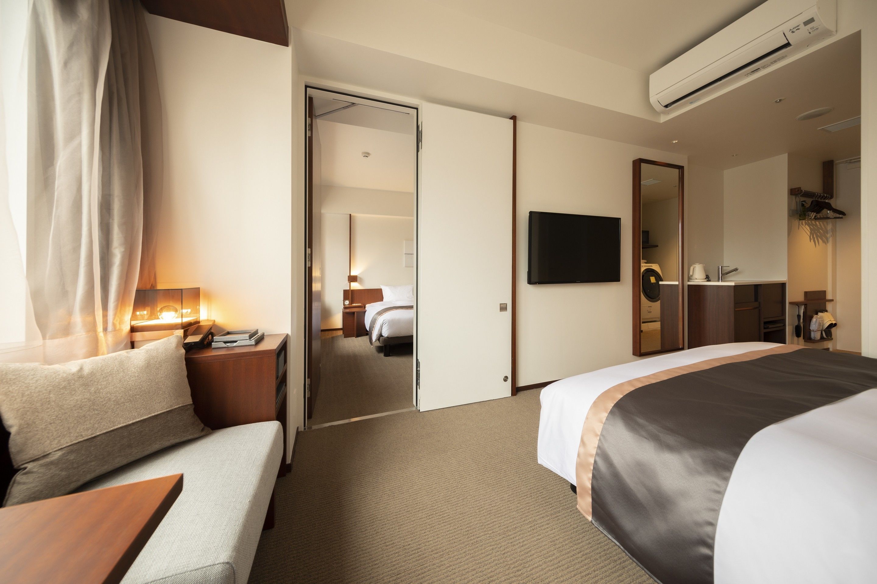 Habitación de hotel con cama, sofá, televisión, espejo y baño visible a través de una puerta abierta.