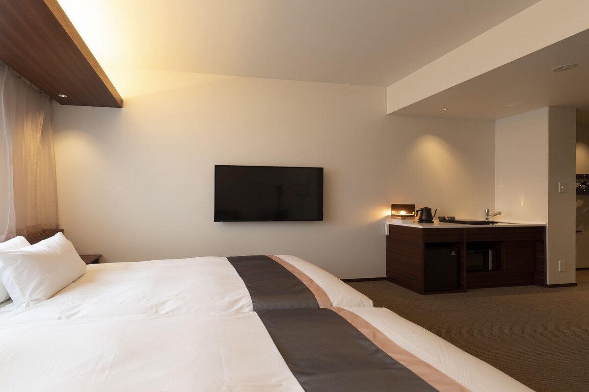A hotel room with a bed, a TV mounted on the wall, a bedside table with a lamp, and a view of the outside through a window.