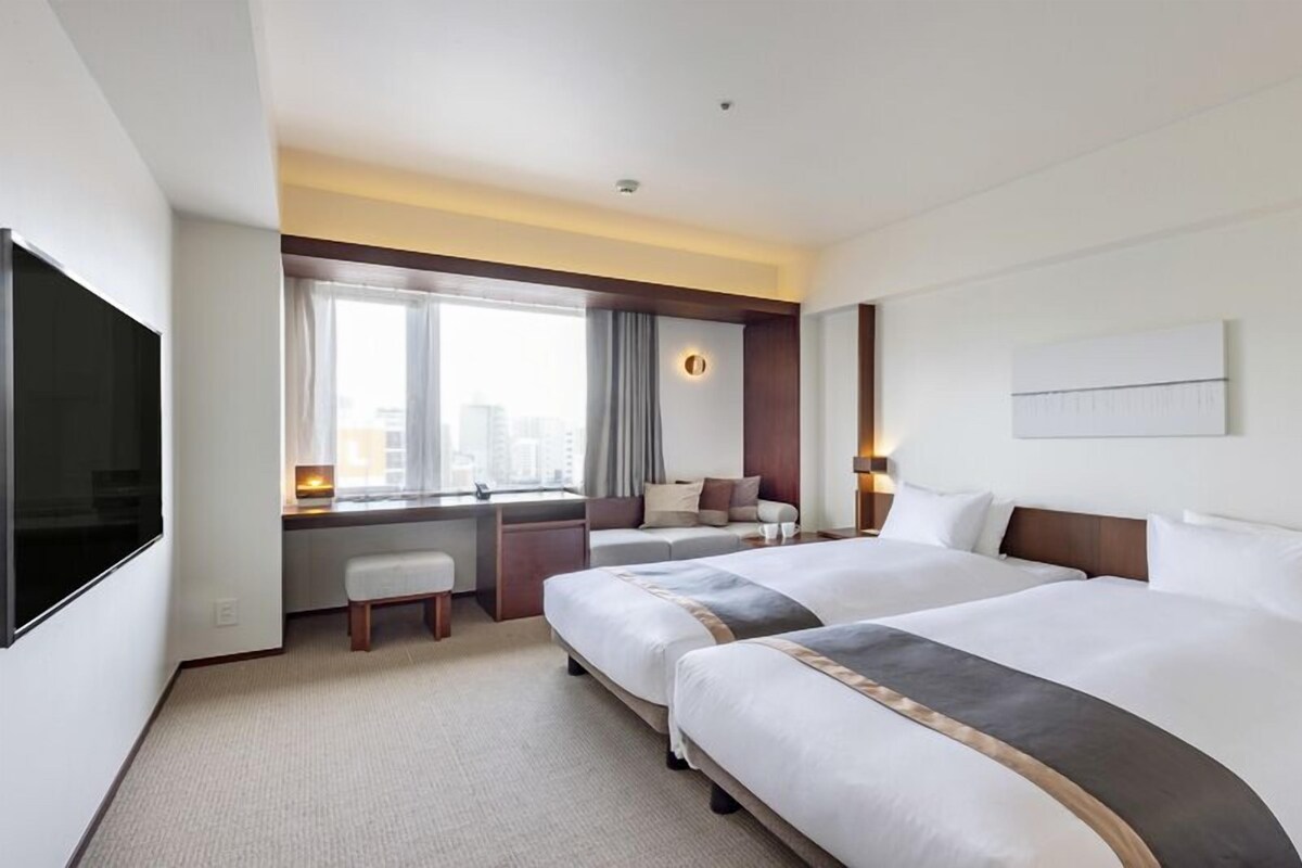 A hotel room with two beds, a desk, a TV, and a city view.