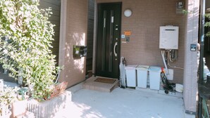 Entrada de edificio con pared de ladrillo, puerta negra, un escalón y zona de servicios con calentador de agua y contenedores de basura.
