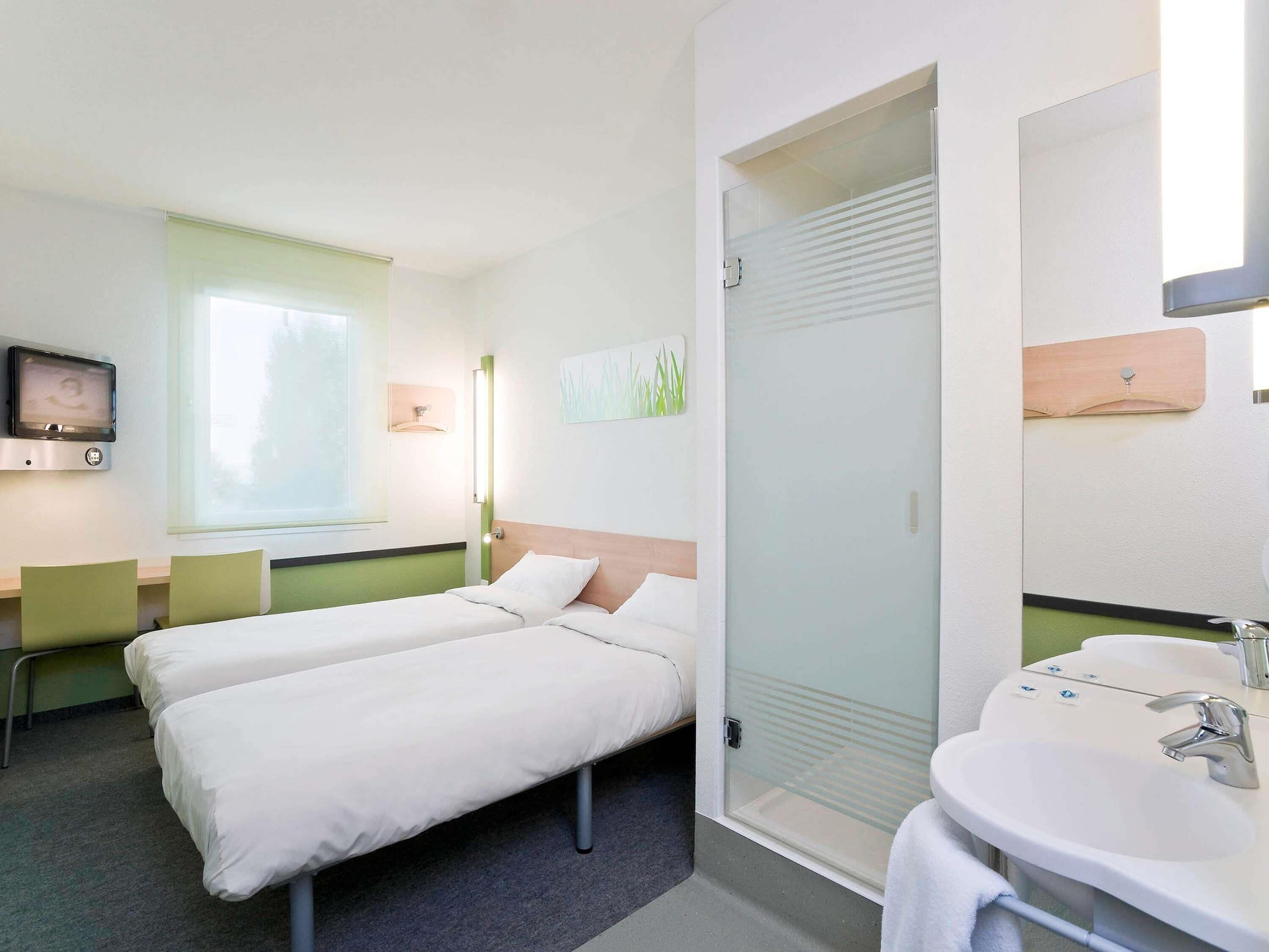 A hotel room with two beds, a TV, a window, a bathroom with a sink and mirror, and a door with a frosted glass panel.