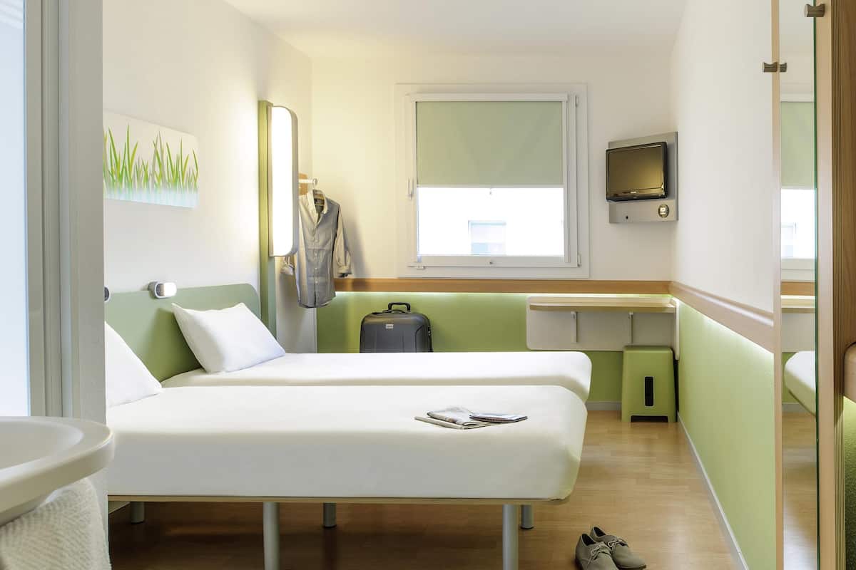 A hotel room with two beds, a desk, a TV, and a window.