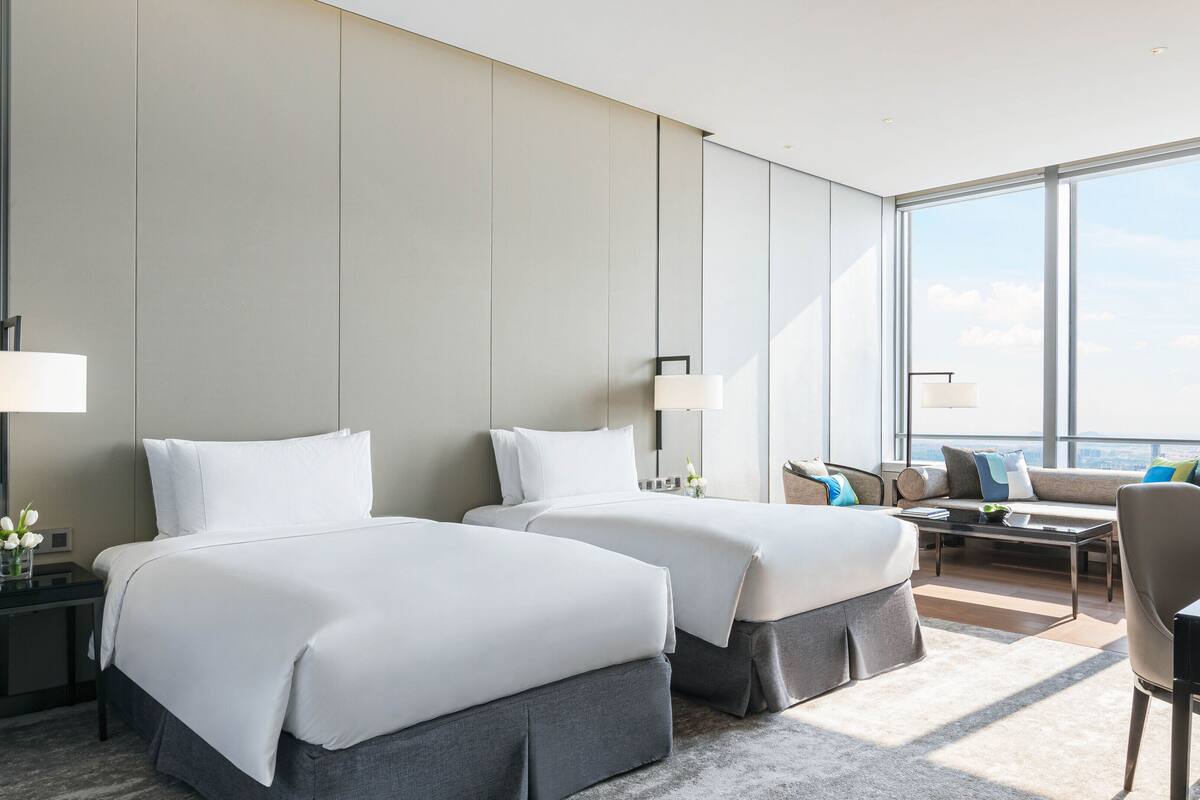 A modern hotel room with two beds, a sofa, a coffee table, and a view of the city.