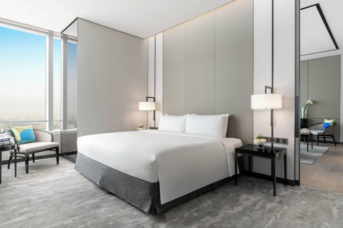 A modern hotel room with a large bed, two bedside tables with lamps, a chair, and a view of the city through large windows.