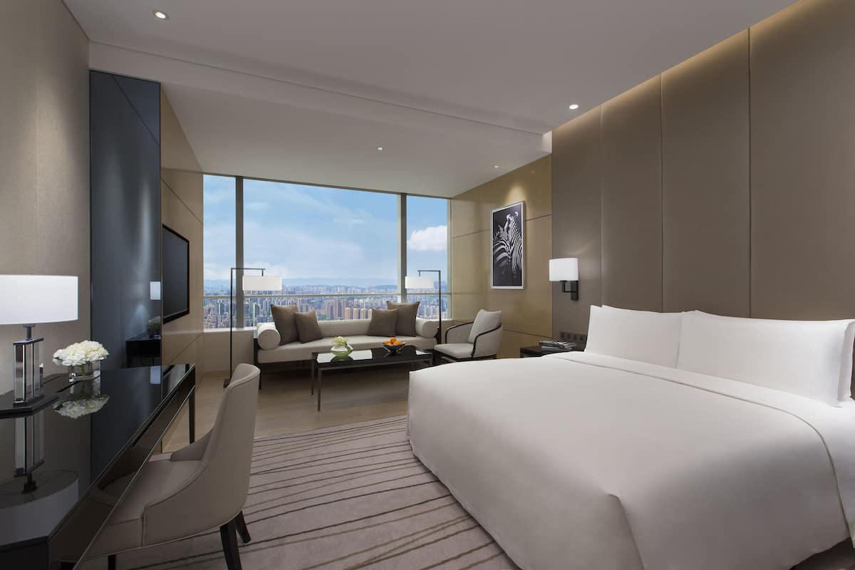 A modern hotel room with a large bed, a sofa, a desk, and a view of the city.