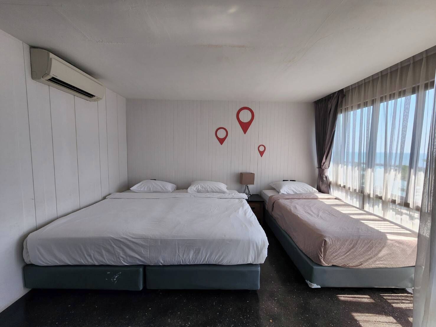 A hotel room with two beds, an air conditioning unit, a nightstand, and a view of the sea through sheer curtains.
