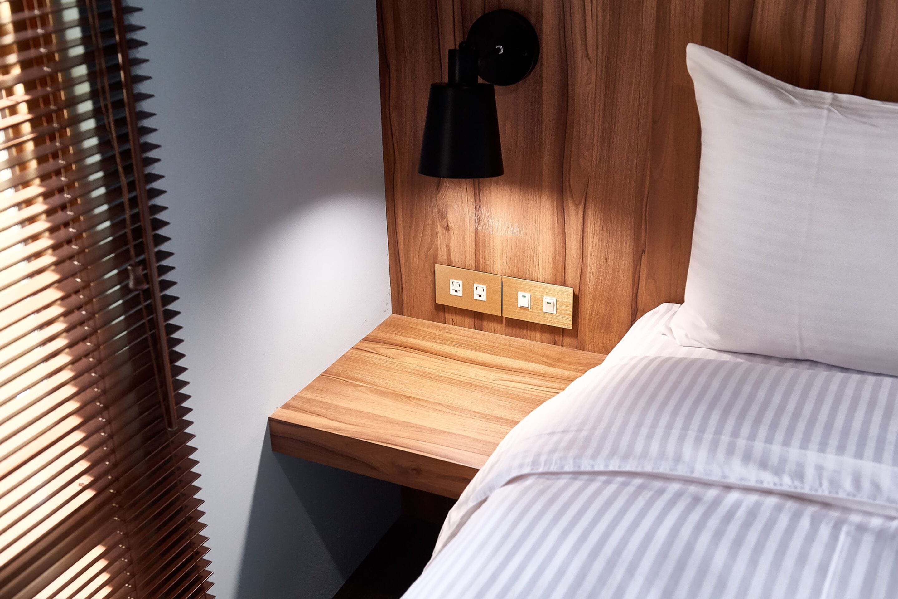 A neatly made bed with white bedding, a wooden nightstand with a black wall-mounted lamp, and a power outlet.