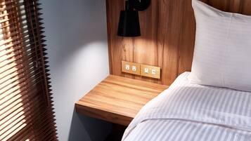 A neatly made bed with white bedding, a wooden nightstand with a black wall-mounted lamp, and a power outlet.