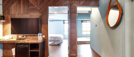 A modern hotel room with a wooden floor, a desk with a chair, a small cabinet, a bed, and a large mirror on the wall.