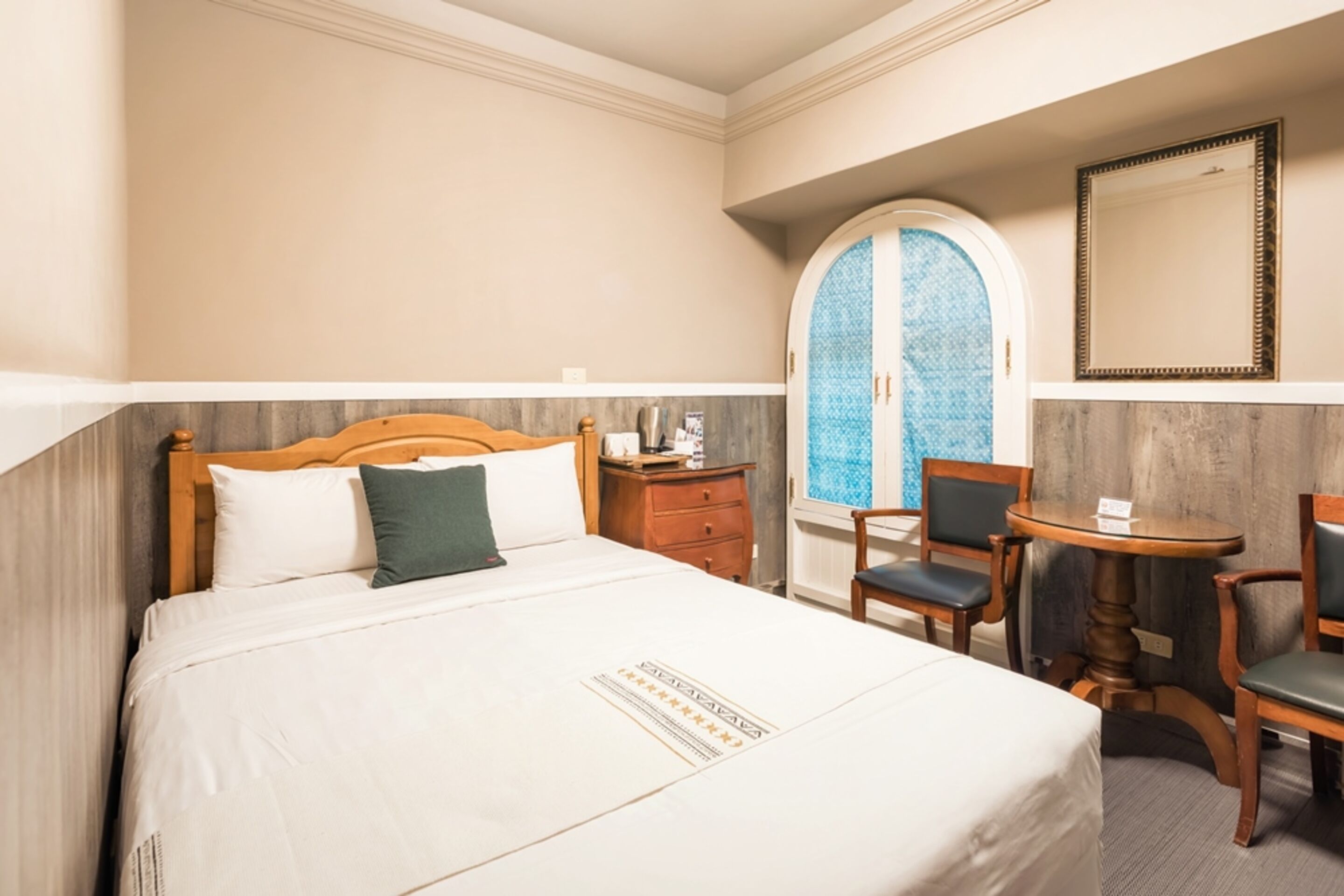 Une chambre d’hôtel avec un lit, une tête de lit en bois, une petite table en bois, deux chaises, une fenêtre donnant sur un paysage agréable et un miroir au mur.