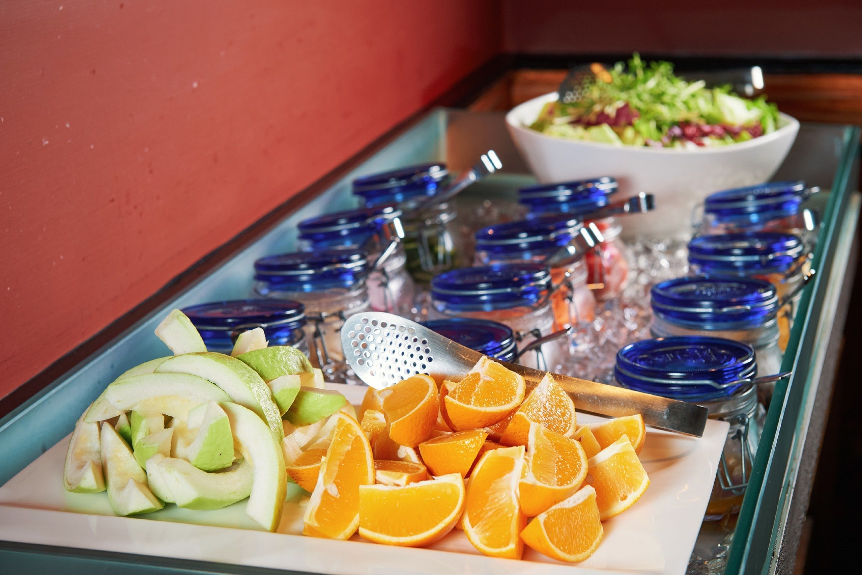 Una mesa de buffet con manzanas en rodajas, naranjas y una ensalada al fondo.