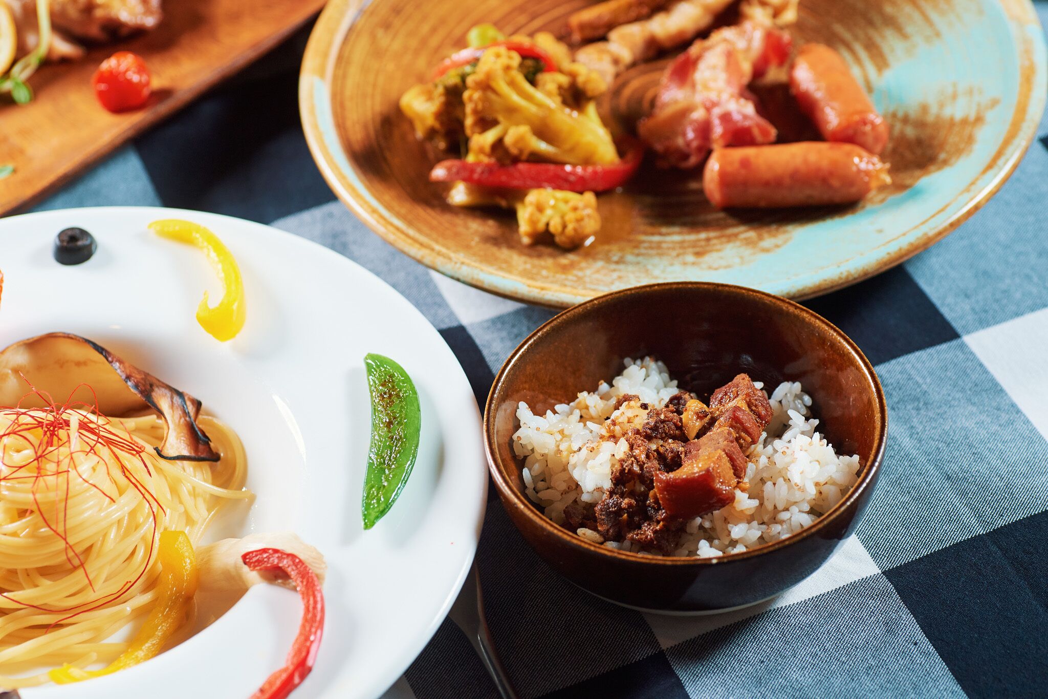 Una mesa con tres platos: fideos y verduras, un bol de arroz con carne, y una fuente de salchichas y verduras.