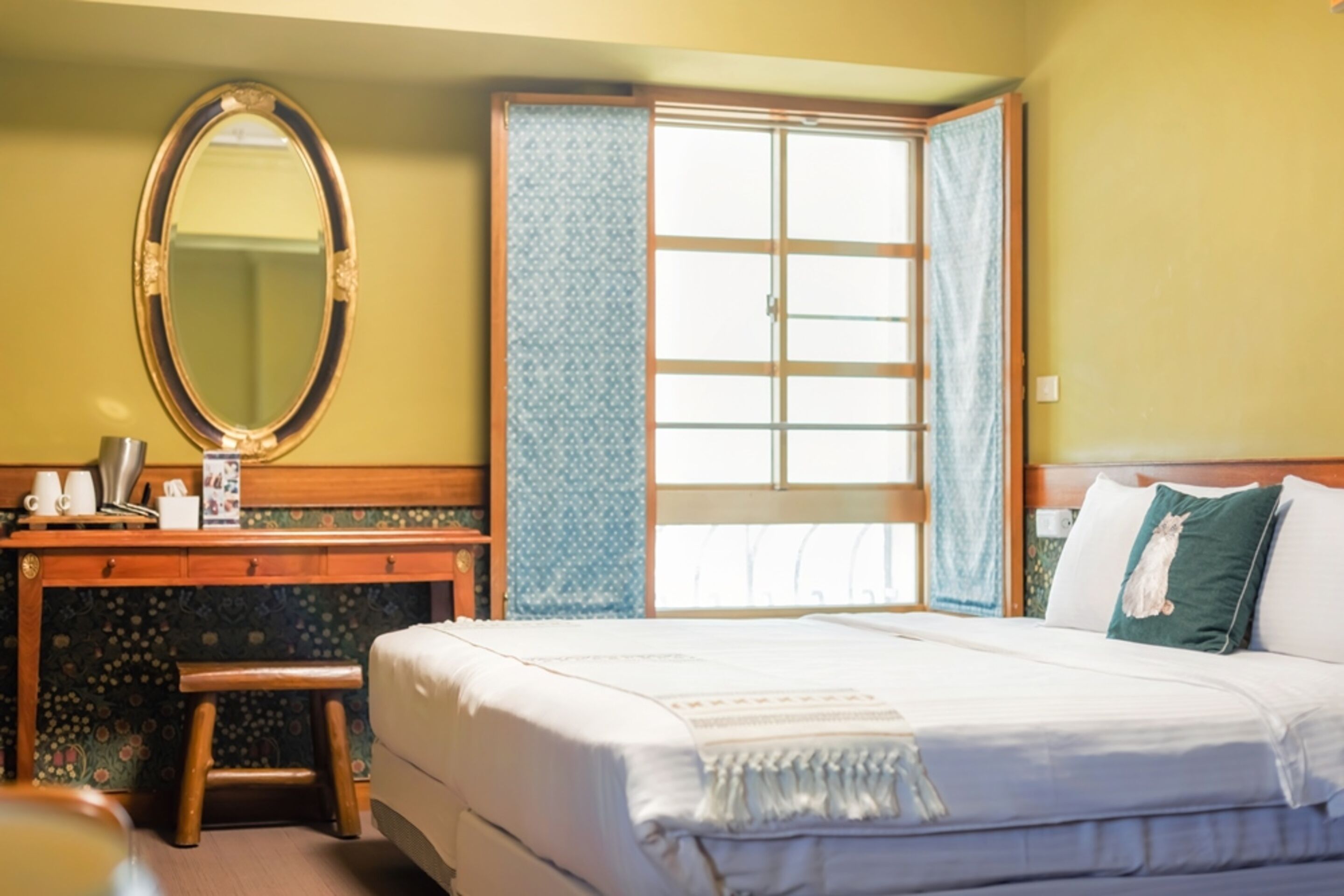Habitación de hotel con cama, escritorio con espejo, taburete pequeño y ventana con cortinas.