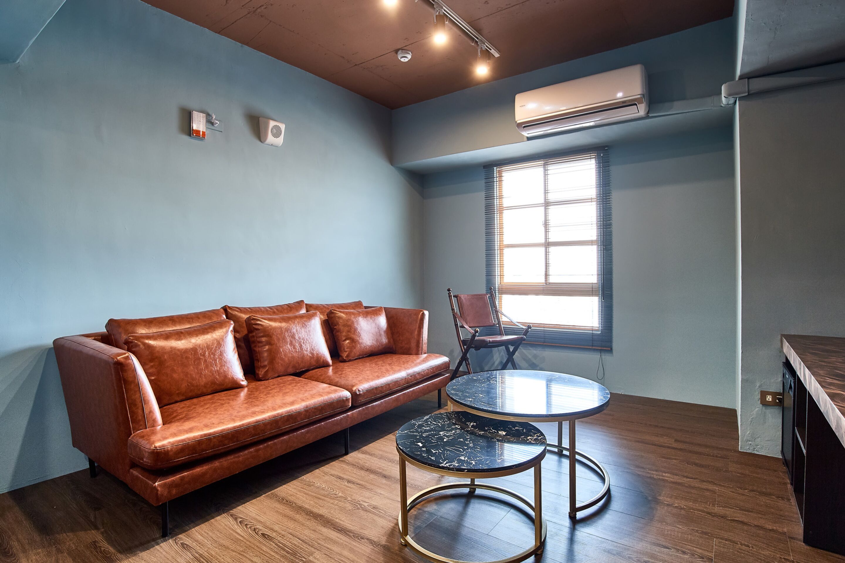 A modern living room with a brown leather sofa, two round tables, a window with blinds, and an air conditioning unit on the wall.