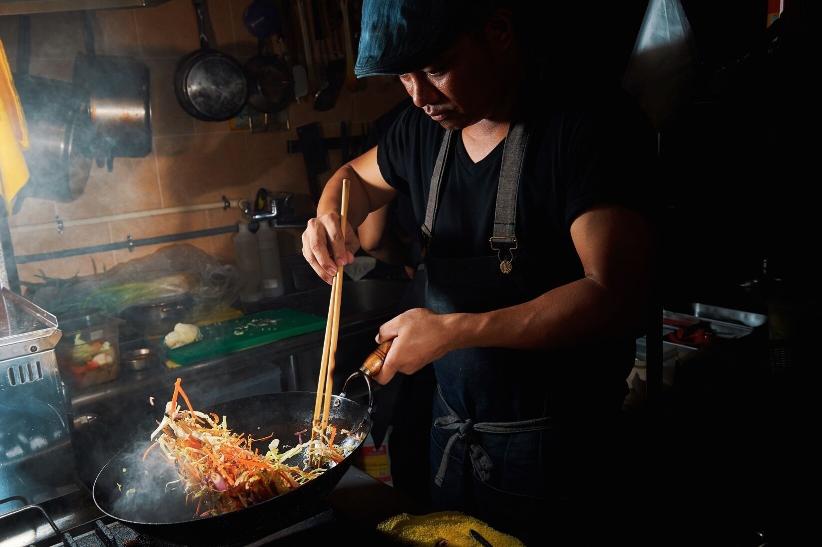 A person cooking in a kitchen with a wok on a stove, a cutting board, and various kitchen utensils.