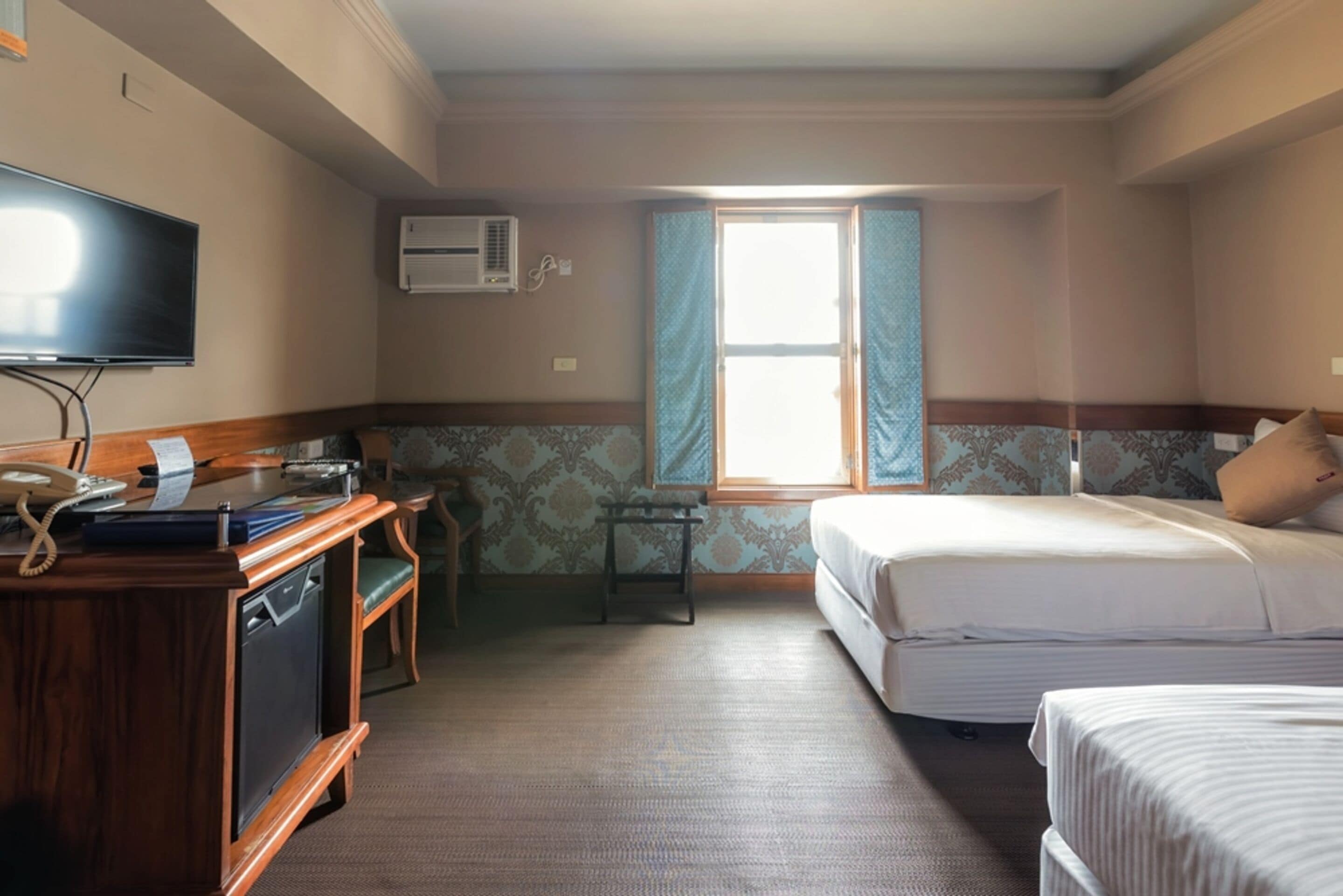 A hotel room with a wooden desk, a television, a bed, and a window with curtains.