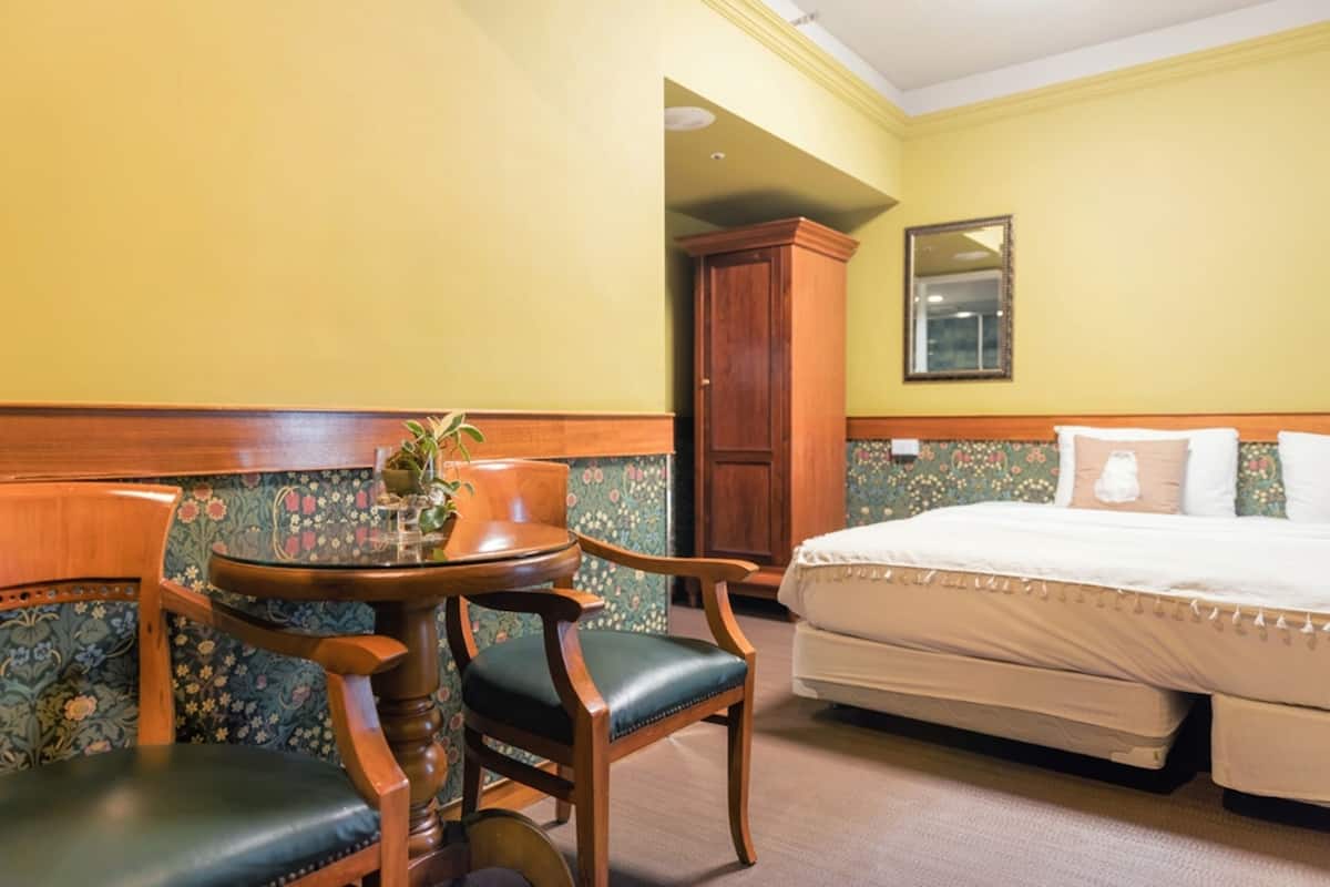 Ein Hotelzimmer mit einem Bett, einem Holzstuhl, einem kleinen runden Tisch mit einer Pflanze und blumigem Wandpapier.