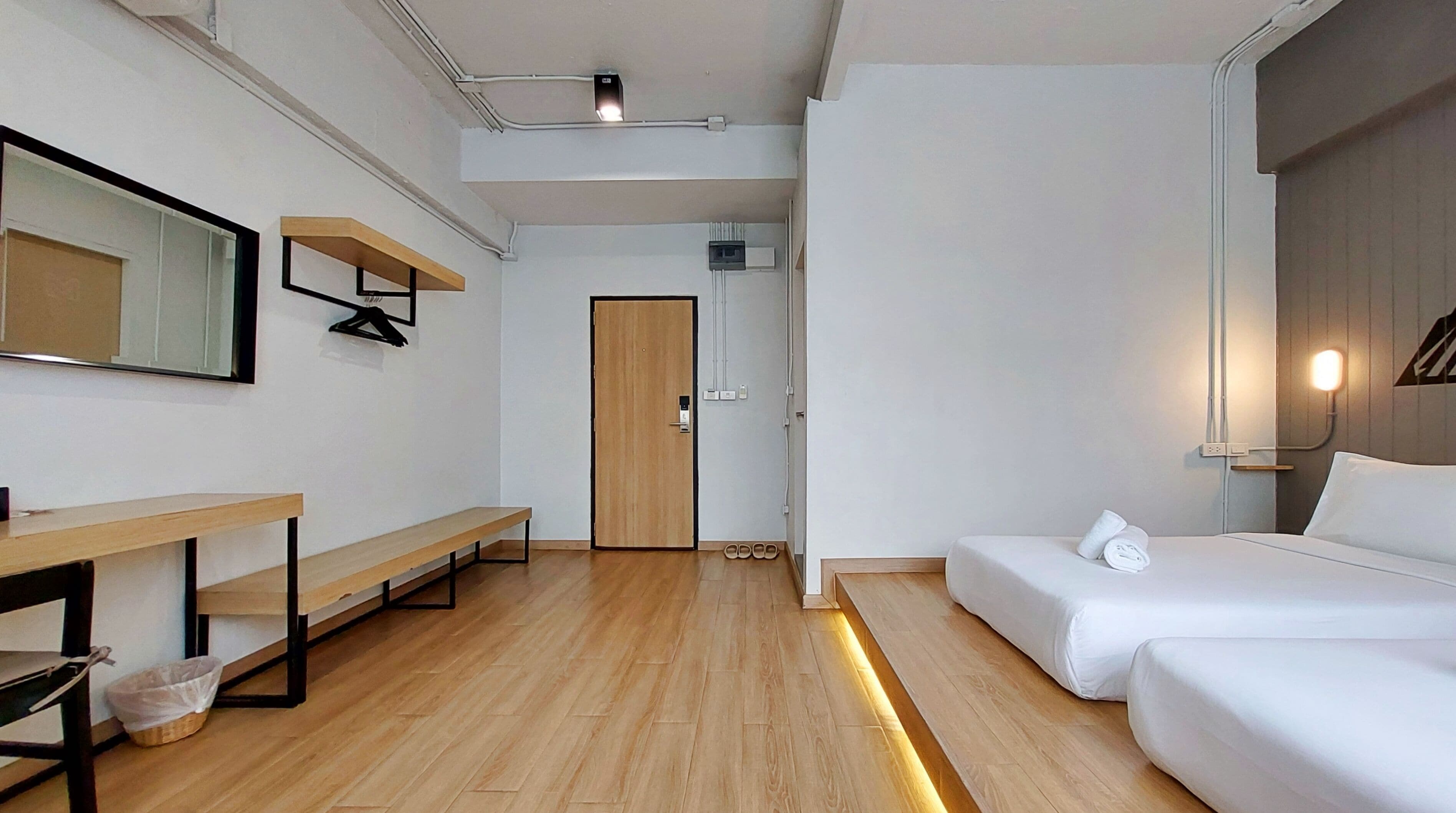 Una habitación de hotel con piso de madera, una cama, un escritorio con silla, un banco, un espejo y una puerta.
