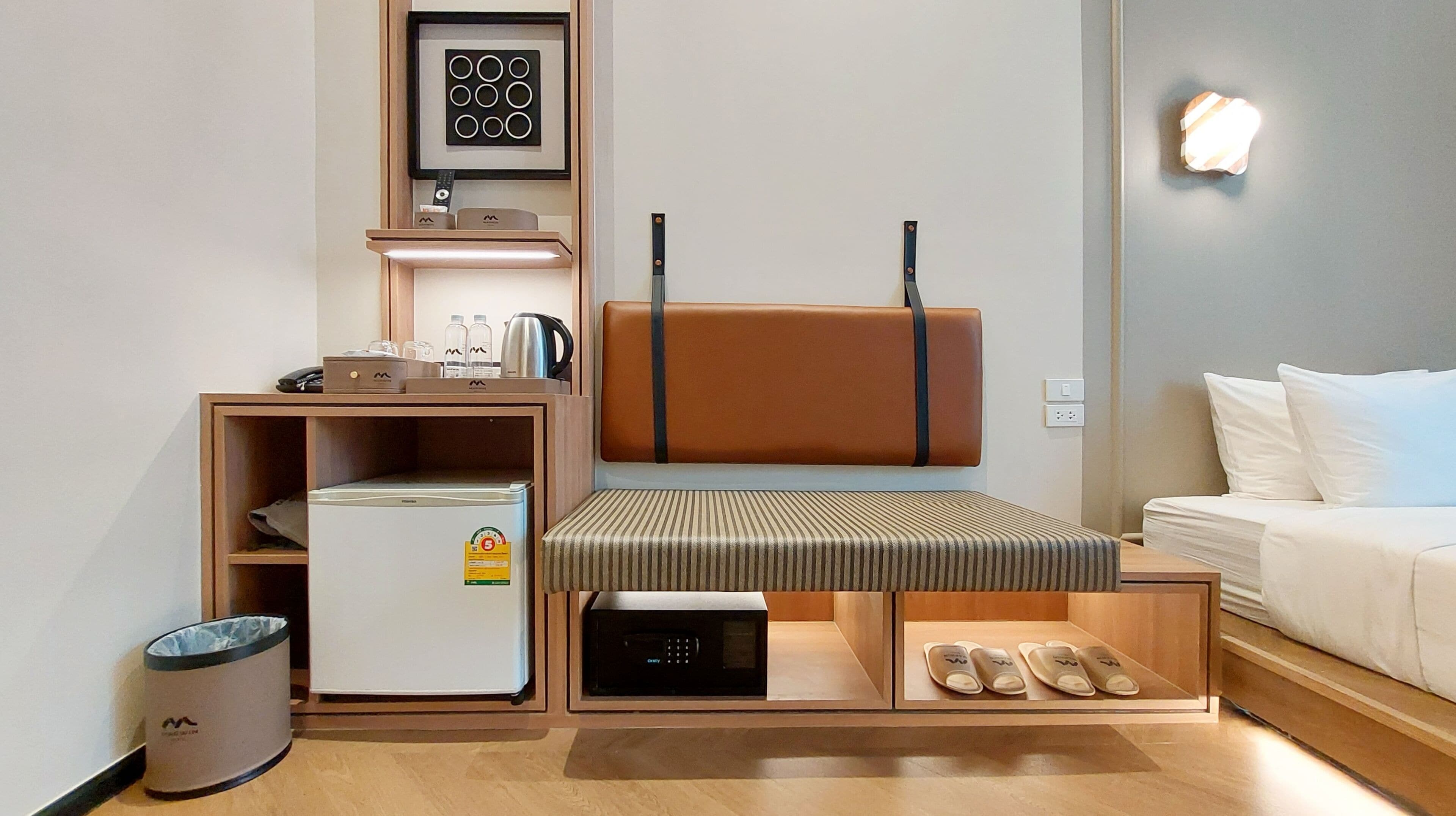 A compact hotel room with a bed, a small kitchenette, a microwave, a trash bin, a shelf with toiletries, and a wall-mounted light.