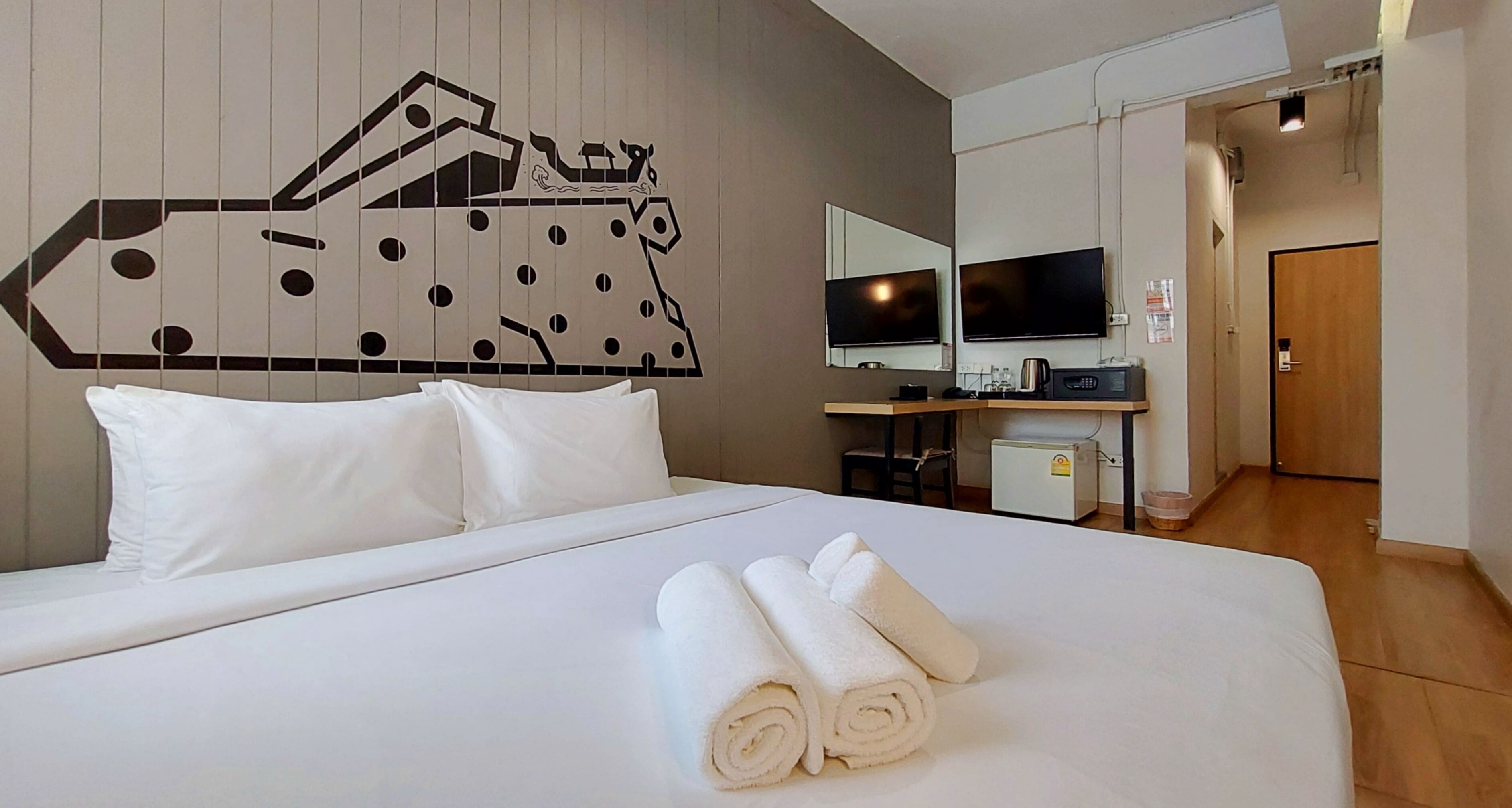 A hotel room with a bed, two pillows, a wall-mounted TV, a small desk with a computer, and a door leading to another room.