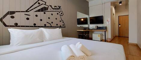 A hotel room with a bed, two pillows, a wall-mounted TV, a small desk with a computer, and a door leading to another room.