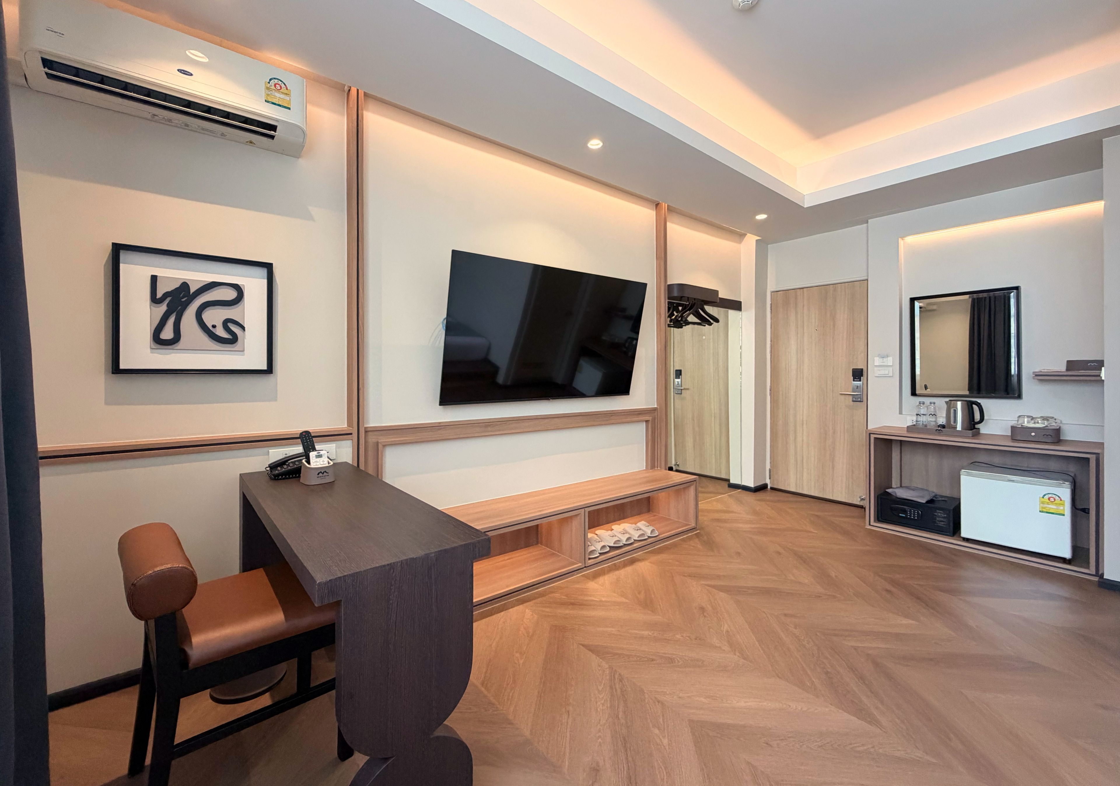 Una habitación de hotel moderna con televisor de pantalla plana, un escritorio con silla, una kitchenette pequeña y un espejo.