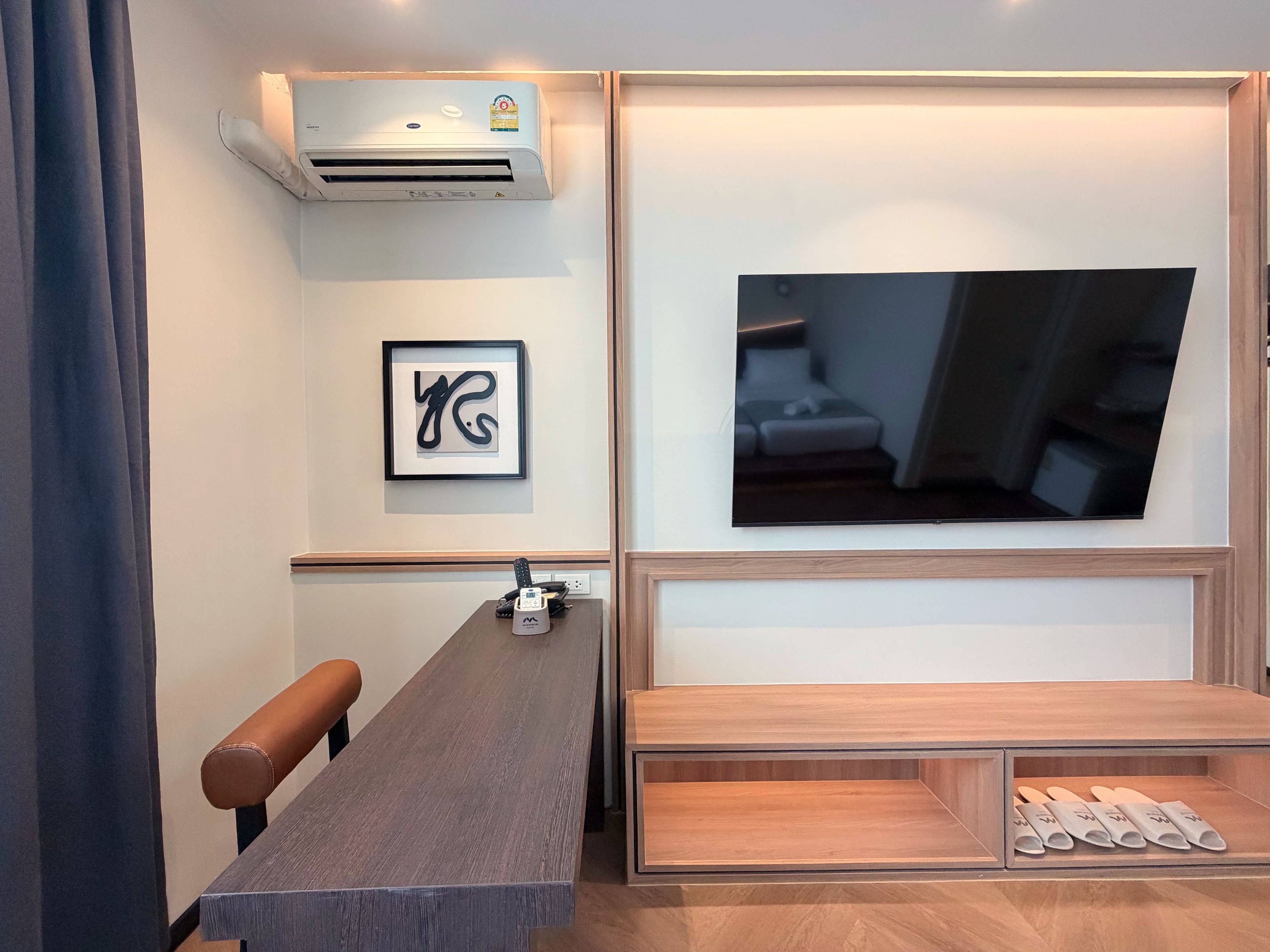 A modern room with a wooden table, a mounted air conditioner, a framed artwork, a television, and a bench.