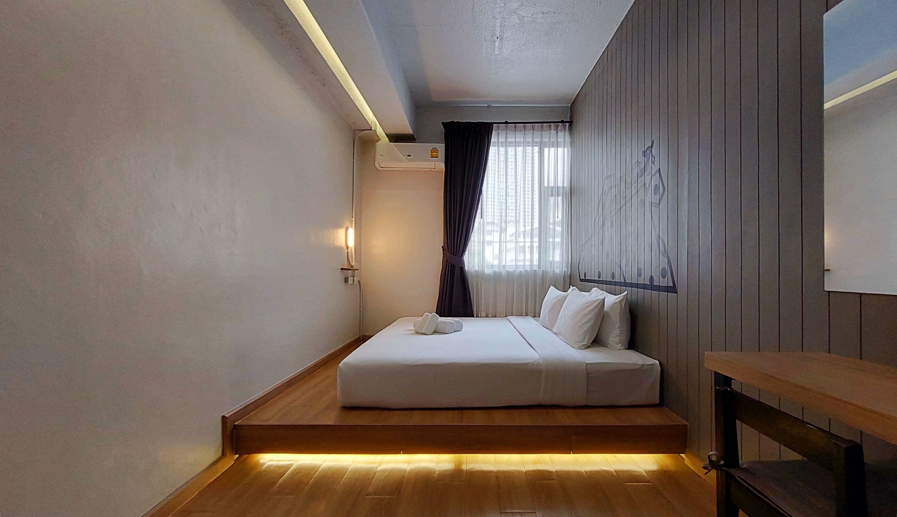 Een hotelkamer met een bed, een houten bankje, een luchtverkoeler aan de muur en een raam met gordijnen.