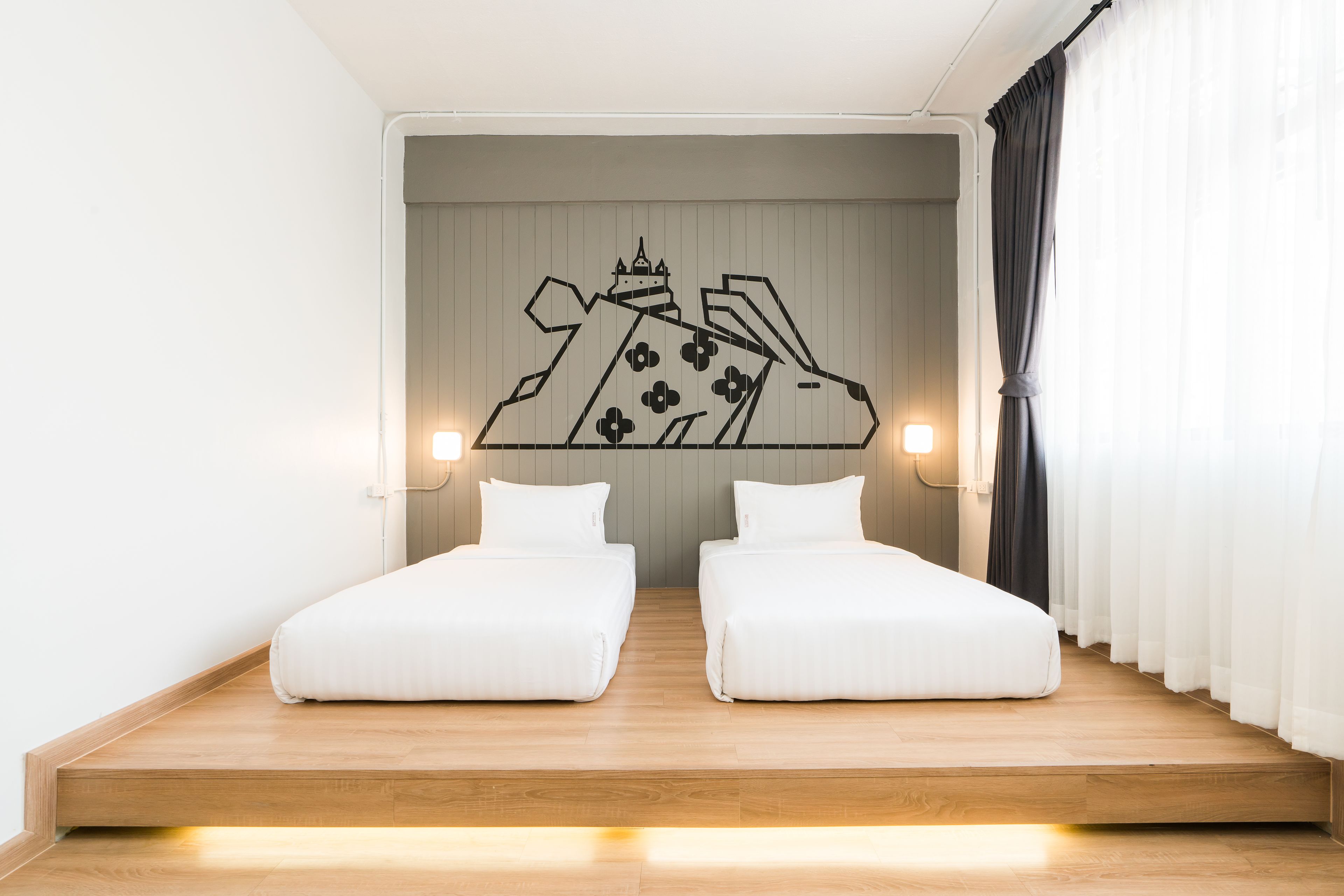 A room with two single beds, a wooden platform, a wall-mounted artwork of a building, and a window with curtains.