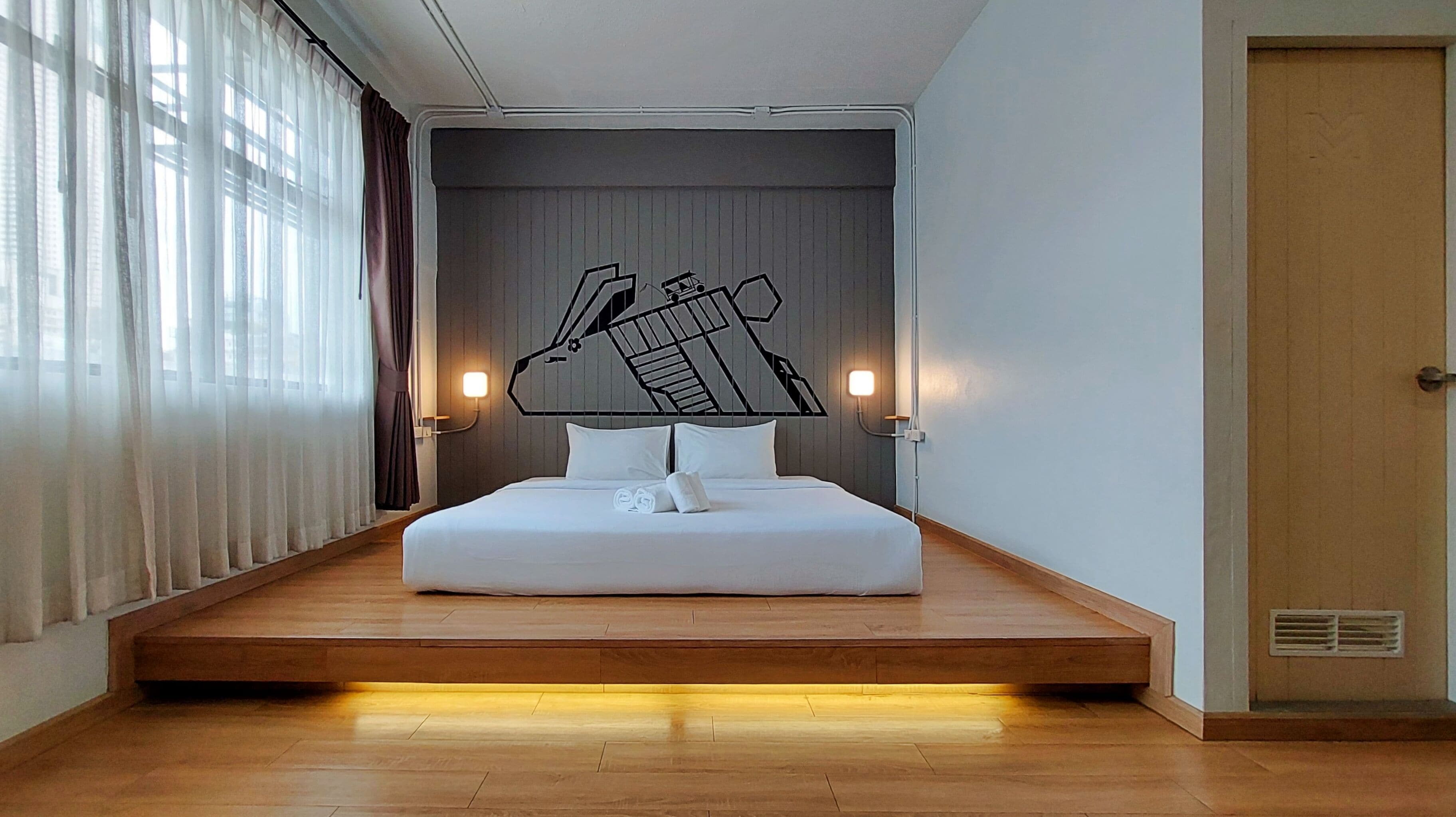 A modern hotel room with a bed centered on a platform, a large window with curtains, and a door on the right.